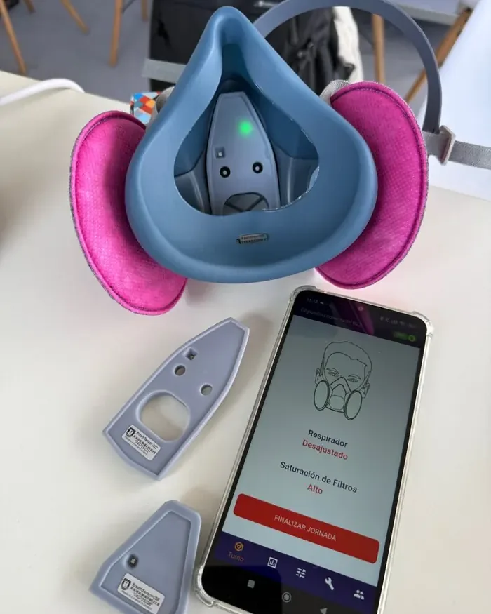 Primer plano del sensor RespiraIA+ instalado en un respirador industrial, junto a la app móvil mostrando el estado de ajuste y saturación de filtros